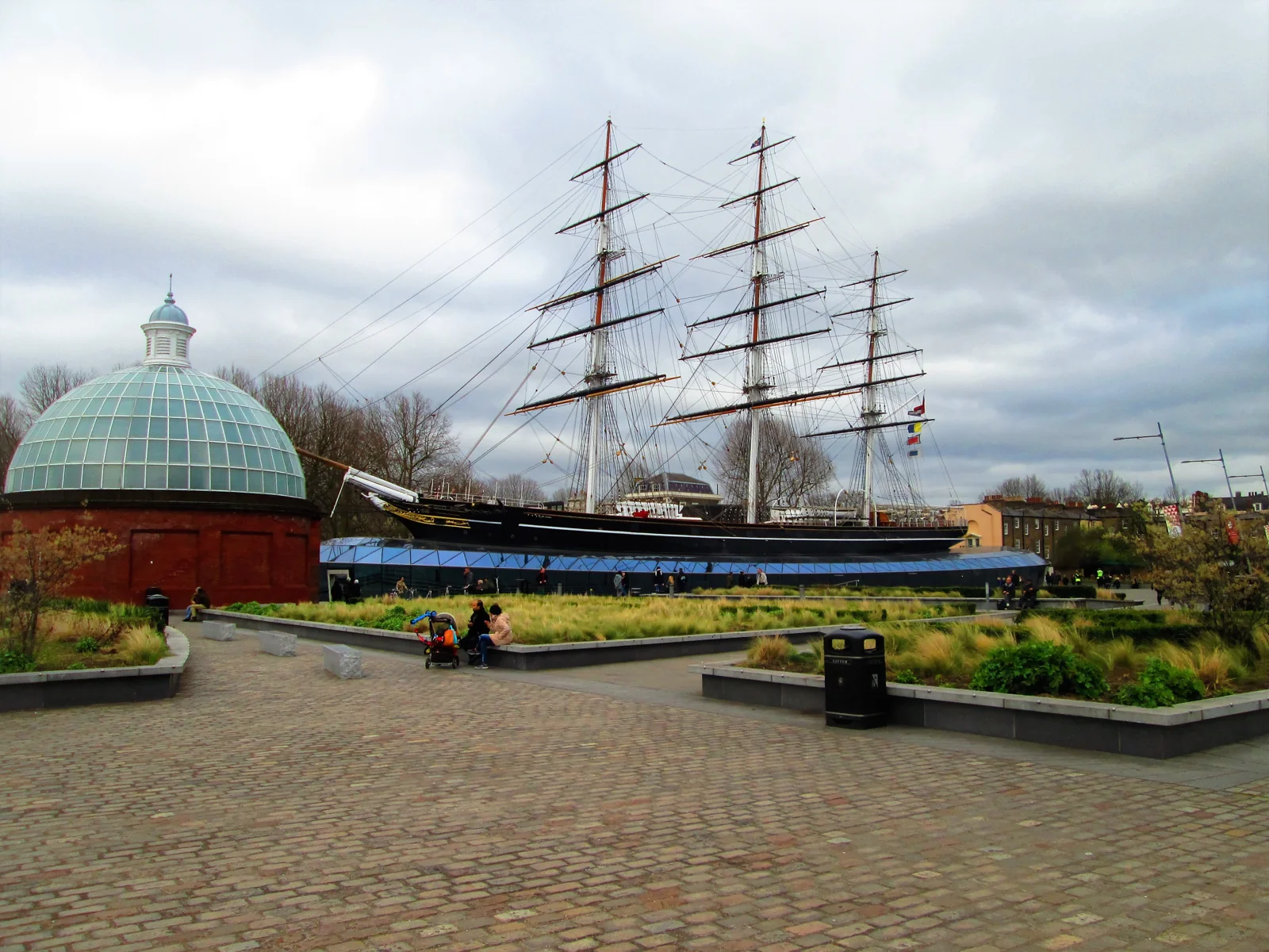Cutty Sark’s three masts and black hull rise above the glass canopy in Greenwich, beside the domed foot tunnel entrance.